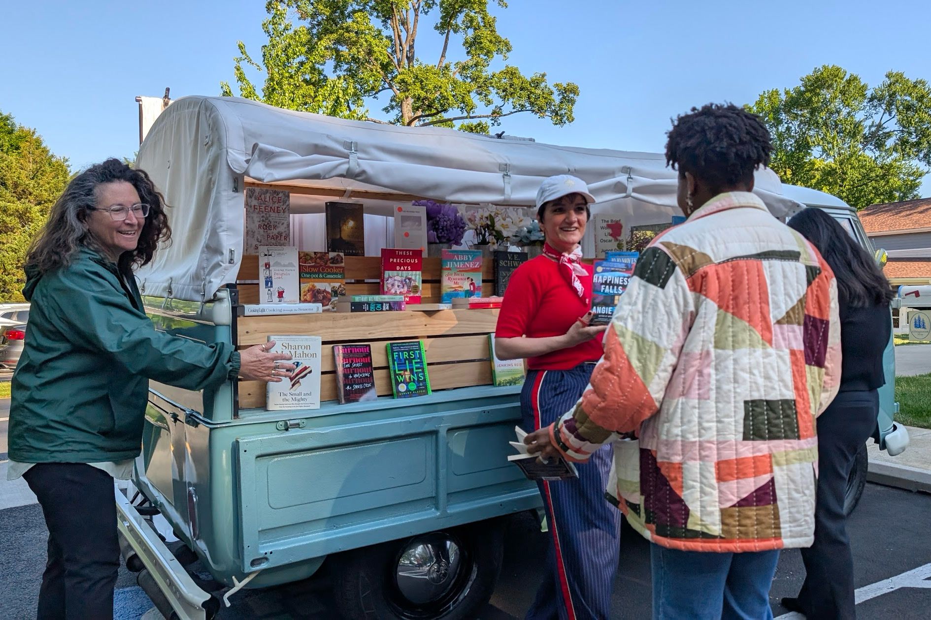 Guests gathered around Rosi's truck bed, fitted with wooden shelves of books under her canvas canopy, as a host in a red top talks with them.