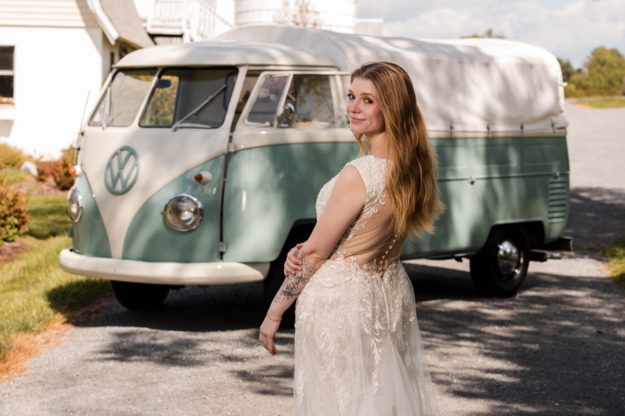 A bride in a lace gown looks back over her shoulder on a sunny lane, with Rosi, a mint-teal and cream vintage Volkswagen pickup, parked behind her.
