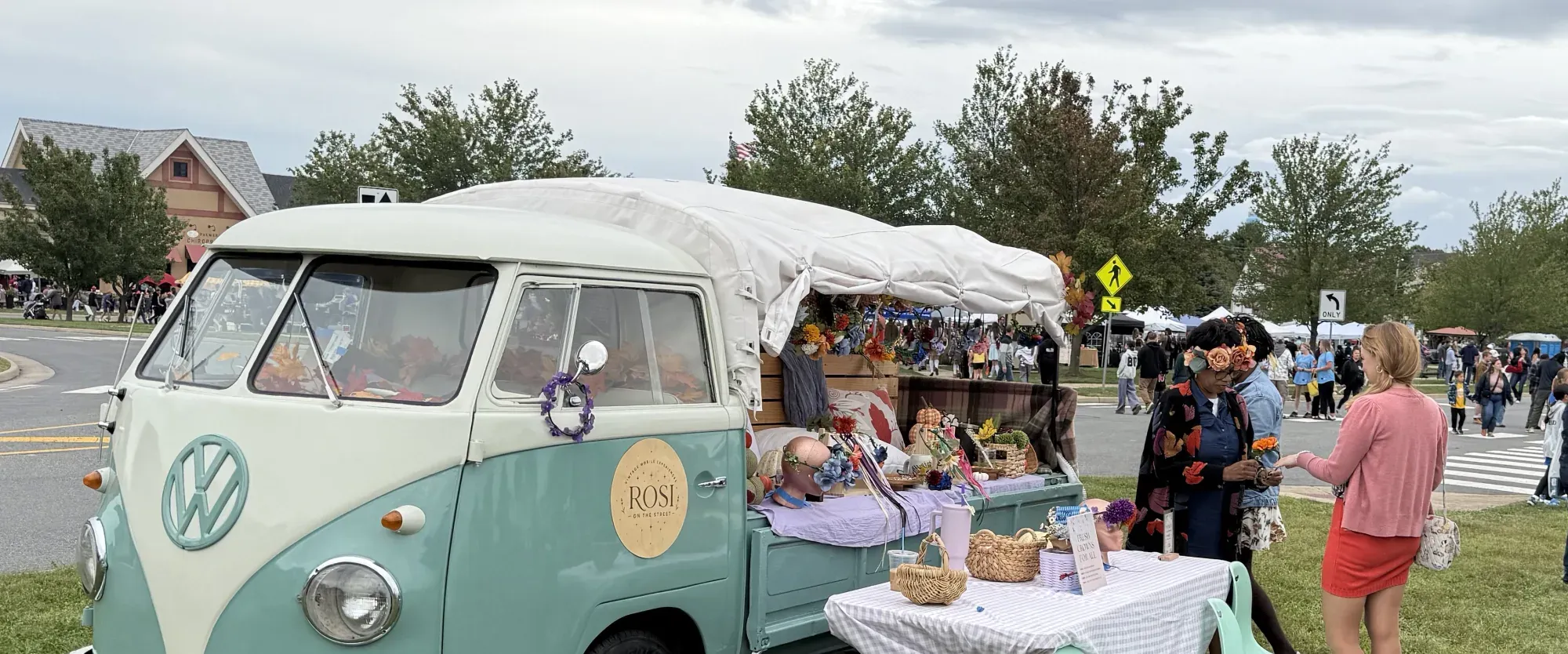 Rosi, a teal and cream 1962 Volkswagen single-cab pickup, parked on grass at an outdoor event, with flower crowns displayed in her bed under a canvas canopy, a gingham-covered table beside her, and two women looking at the flower crowns.