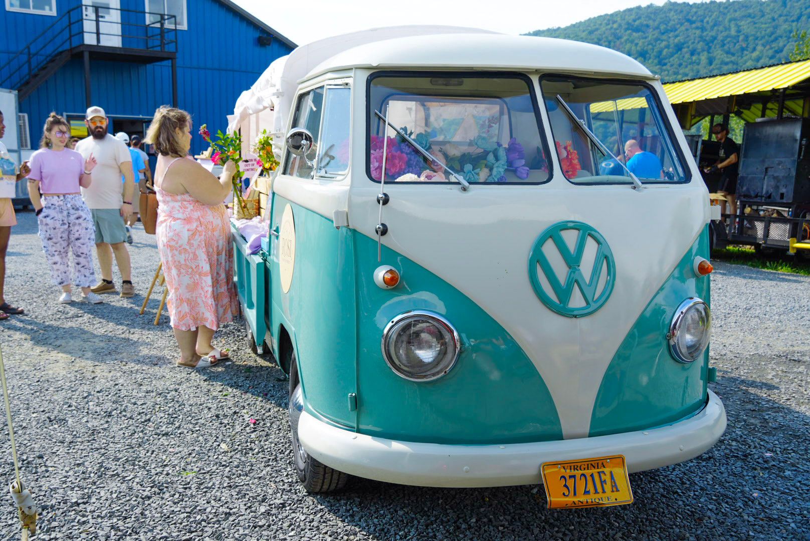 Rosi, a mint-teal and cream 1962 Volkswagen pickup, seen head-on at an outdoor event, with a guest in a floral dress building a bouquet at the flower bar in her bed.