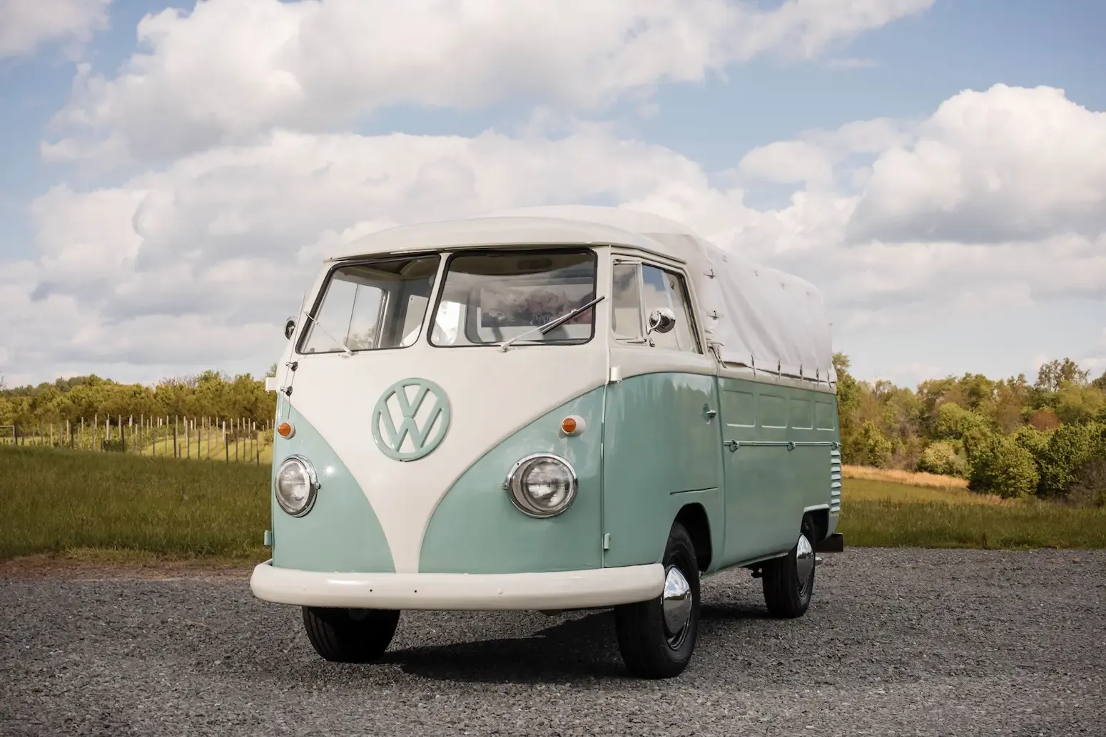 Rosi, a teal and cream 1962 Volkswagen single-cab pickup with a canvas cover over her bed, parked on gravel in front of a vineyard under a cloudy blue sky.