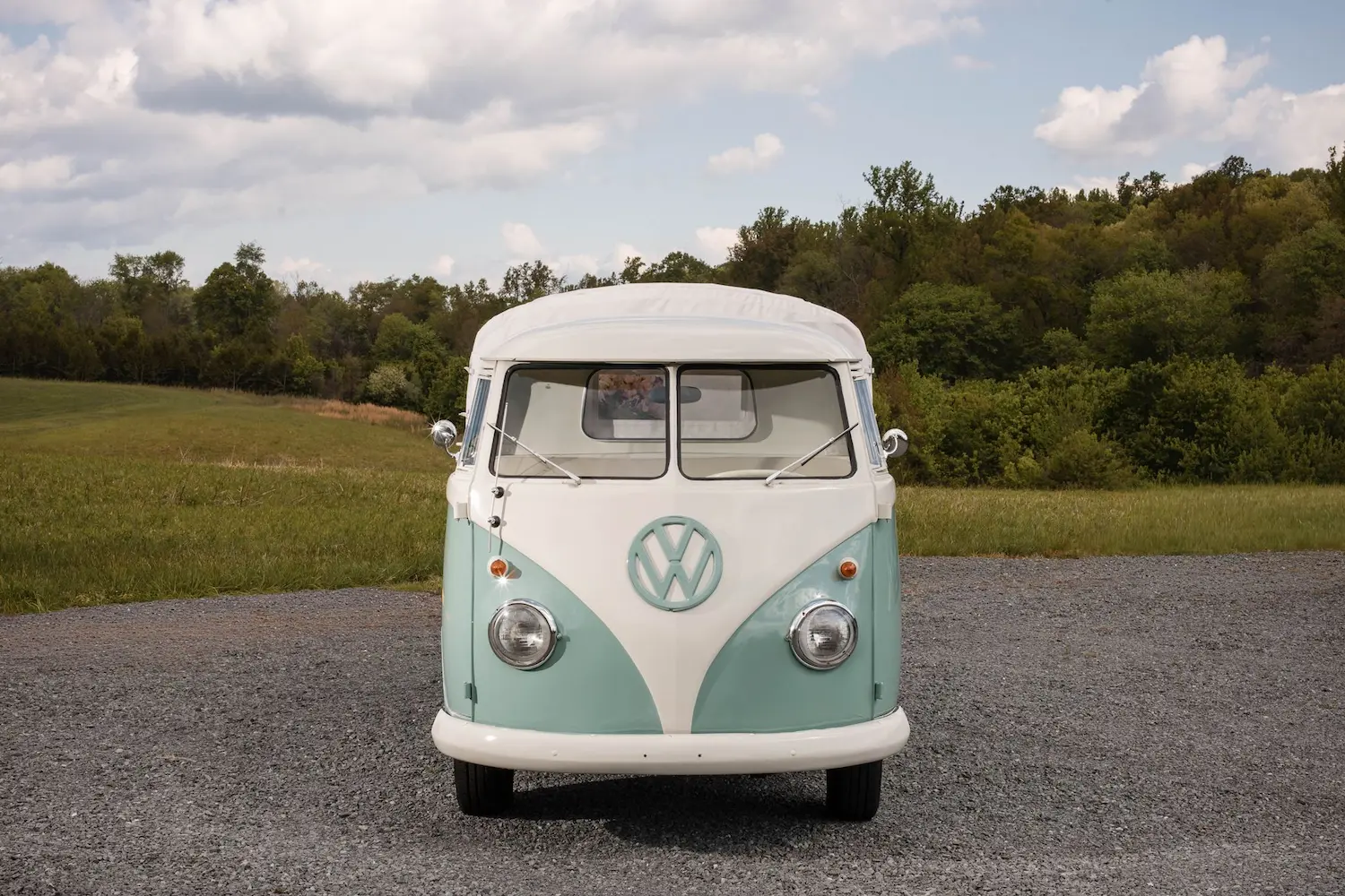 Front view of Rosi, a vintage mint and white VW event truck, parked on a gravel road with trees and open field behind it.